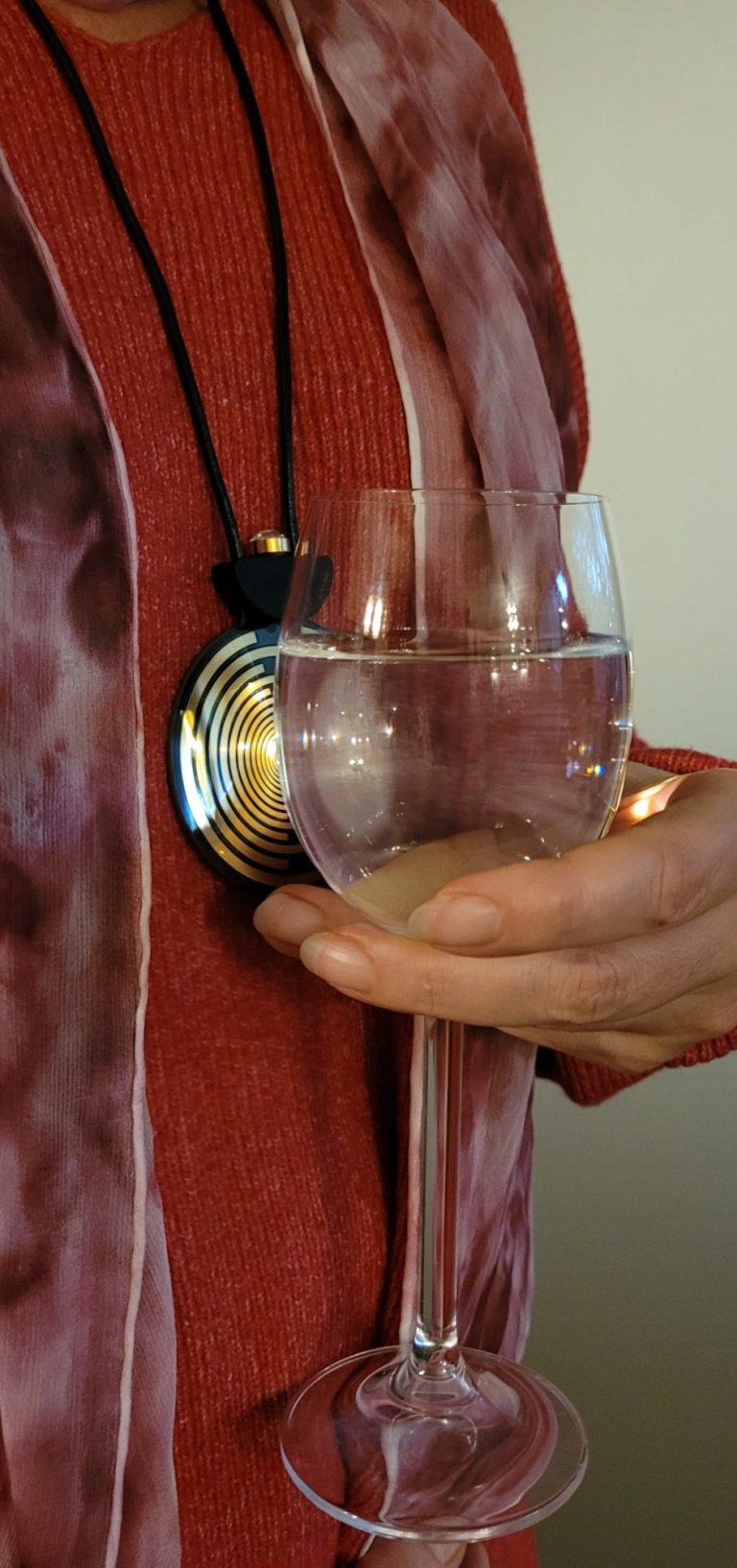 Wasser strukturieren mit Mediator Eine Hand hält ein Weinglas gefüllt mit Wasser direkt an den Mediator. Die Person trägt den Mediator auf Brust.