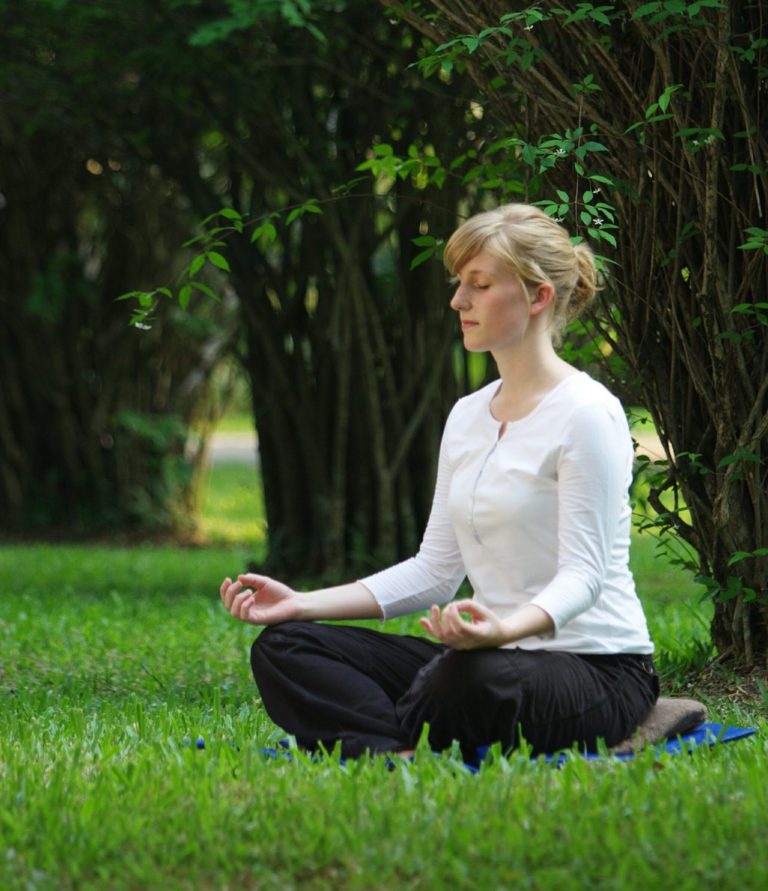 Frau sitzt im Meditationssitz auf dem Gras, umgeben von Bäumen.