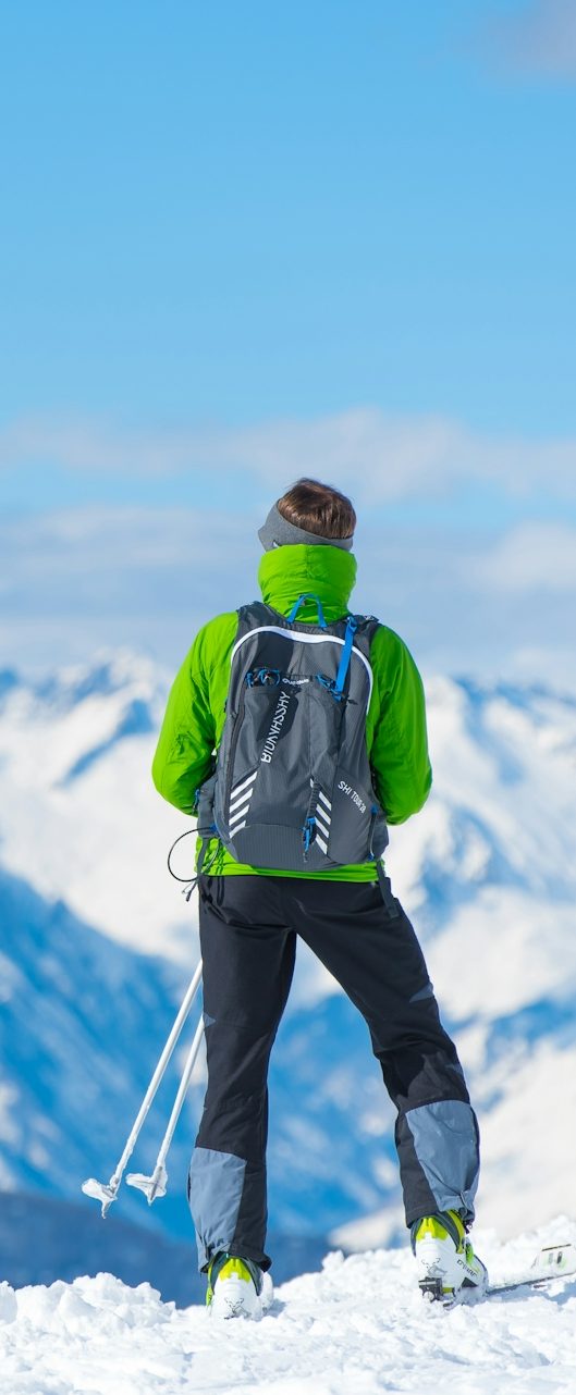 Skifahrer in grüner Jacke und Rucksack, Blick auf verschneite Berge.